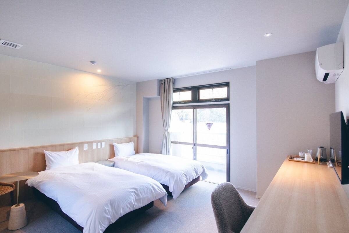 A hotel room with two beds, a desk, a chair, a TV, and a window.