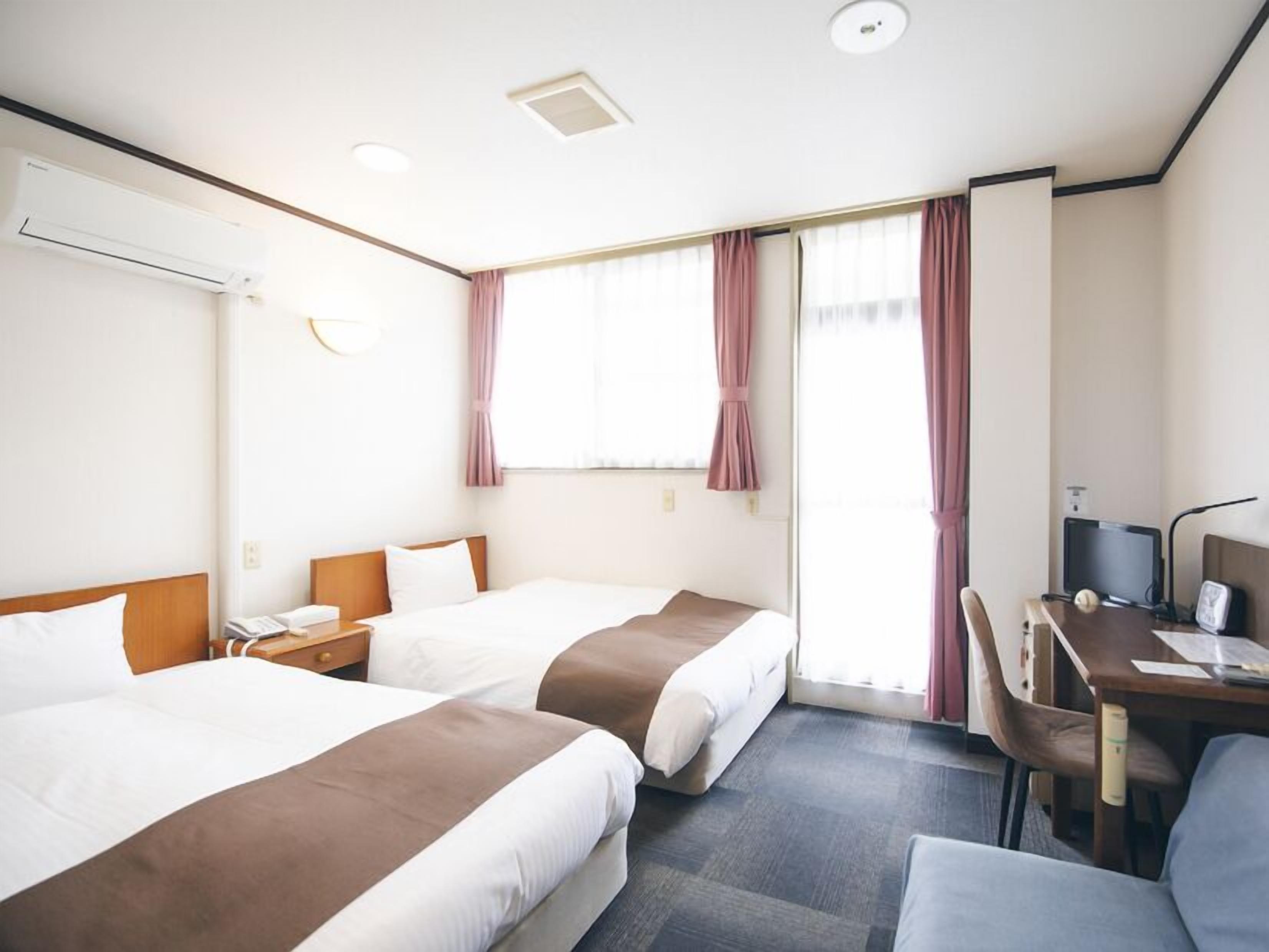 Quarto de hotel com duas camas, uma mesa de escritório, uma cadeira, aparelho de ar condicionado e uma janela com cortinas.