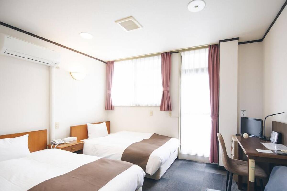 A hotel room with two beds, a desk, a chair, an air conditioning unit, and a window with curtains.