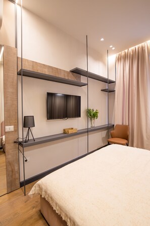 Een moderne slaapkamer met een bed, een televisie aan de muur, een stoel en een klein tafeltje met een lamp en een plant.