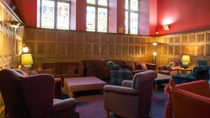 Une bibliothèque aux murs rouges, dotée de grandes fenêtres à vitraux et de boiseries. L’espace détente comprend plusieurs fauteuils et un canapé.