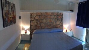 A bedroom with a stone headboard, a bed with a blue bedspread, two bedside lamps, and a painting on the wall. - Lampedusa catatteristic dammuso sea view, 200mt distant, 180° (Lampedusa)