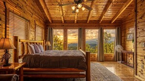 Une chambre située dans un chalet en rondins, avec un lit, des murs en bois, un ventilateur de plafond et de grandes fenêtres offrant une vue sur la montagne.