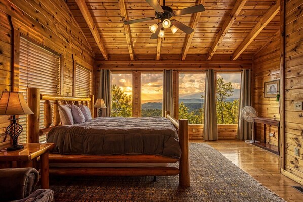 Ein Schlafzimmer in einer Blockhütte, mit einem Bett, Holzwänden, einem Deckenventilator und großen Fenstern mit Blick auf die Berge.