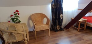 A room with a wooden floor, a wicker chair, a small table, a bed with a red bedspread, a window with curtains, and a potted plant with red flowers. - Conacul Bunicilor (Baia de Fier)