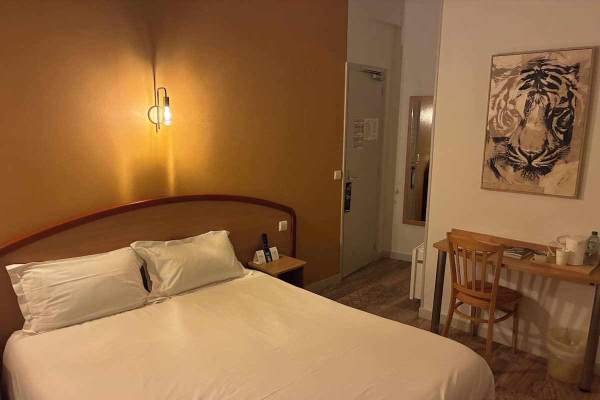 A hotel room with a bed, a desk with a chair, a painting on the wall, and a door leading to another room.
