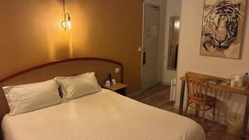 A hotel room with a bed, a desk with a chair, a painting on the wall, and a door leading to another room.