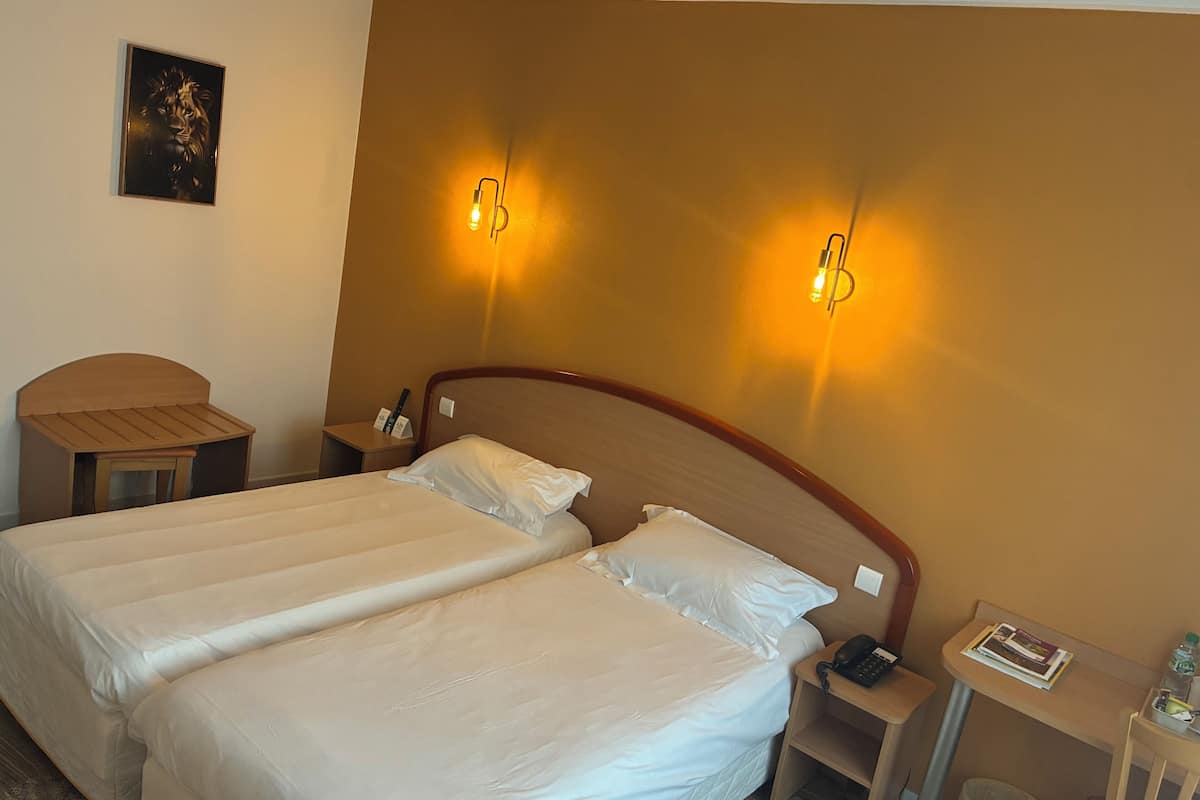 A hotel room with two beds, a wooden headboard, bedside tables with lamps, a framed picture on the wall, and a small table with a phone and a magazine.