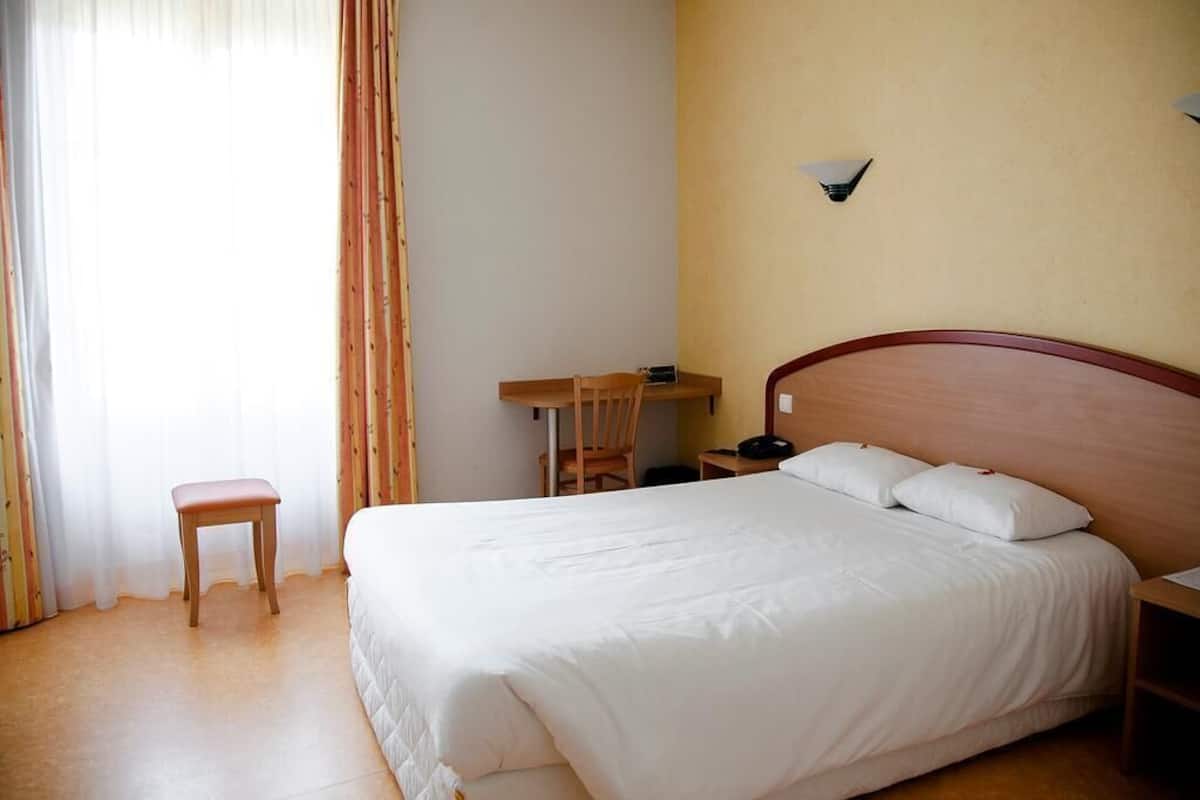 A hotel room with a bed, a desk, a chair, and a small stool.