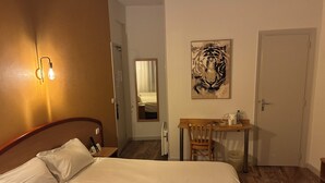 A hotel room with a bed, a desk, a chair, a mirror, and a door. - Hotel du Commerce (Volvic)