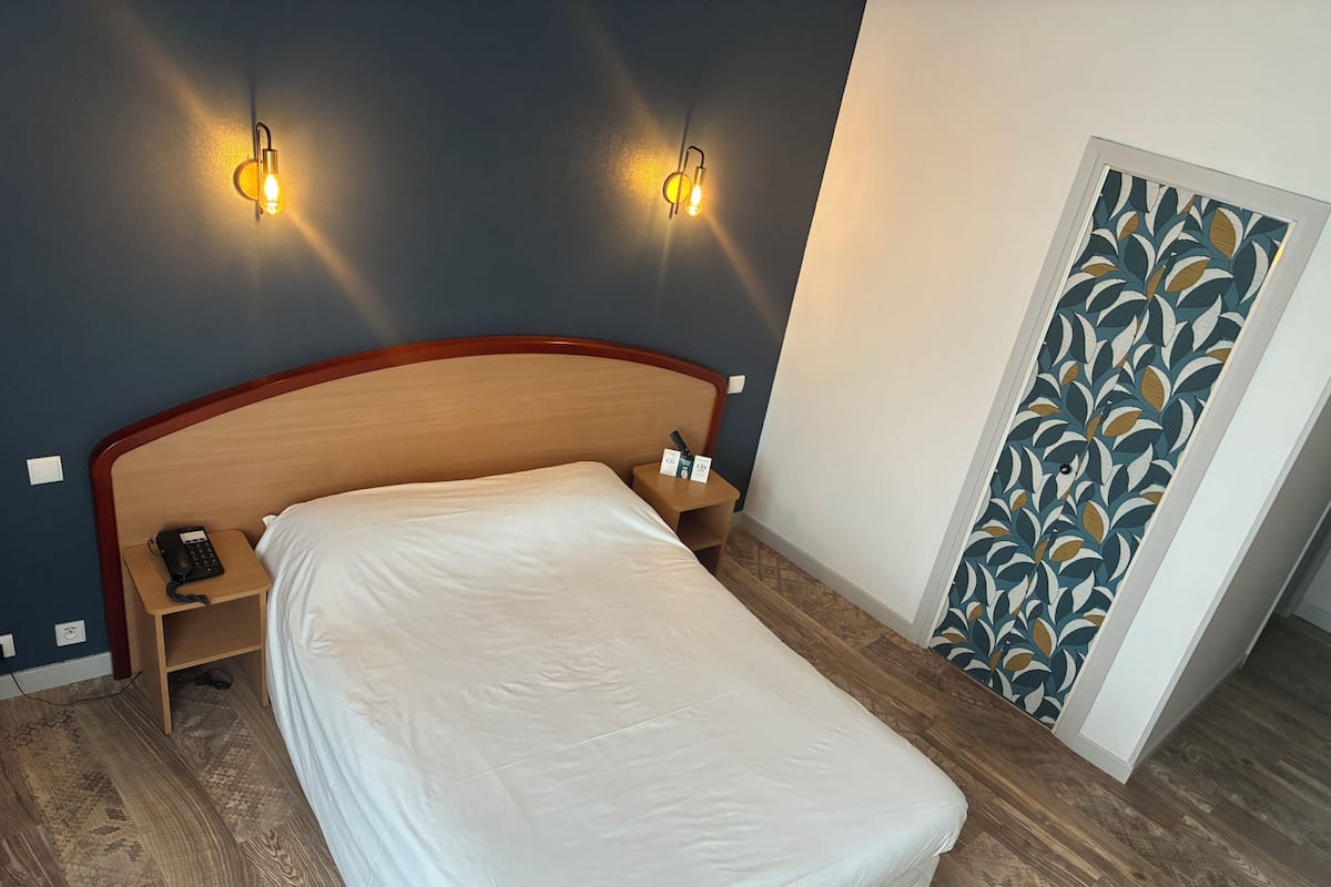 A hotel room with a wooden headboard, a bed with white linens, two bedside tables with lamps, a telephone, and a patterned wall.