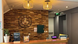 A reception desk with a wooden wall, a large clock, and two pendant lights. - Hotel du Commerce (Volvic)