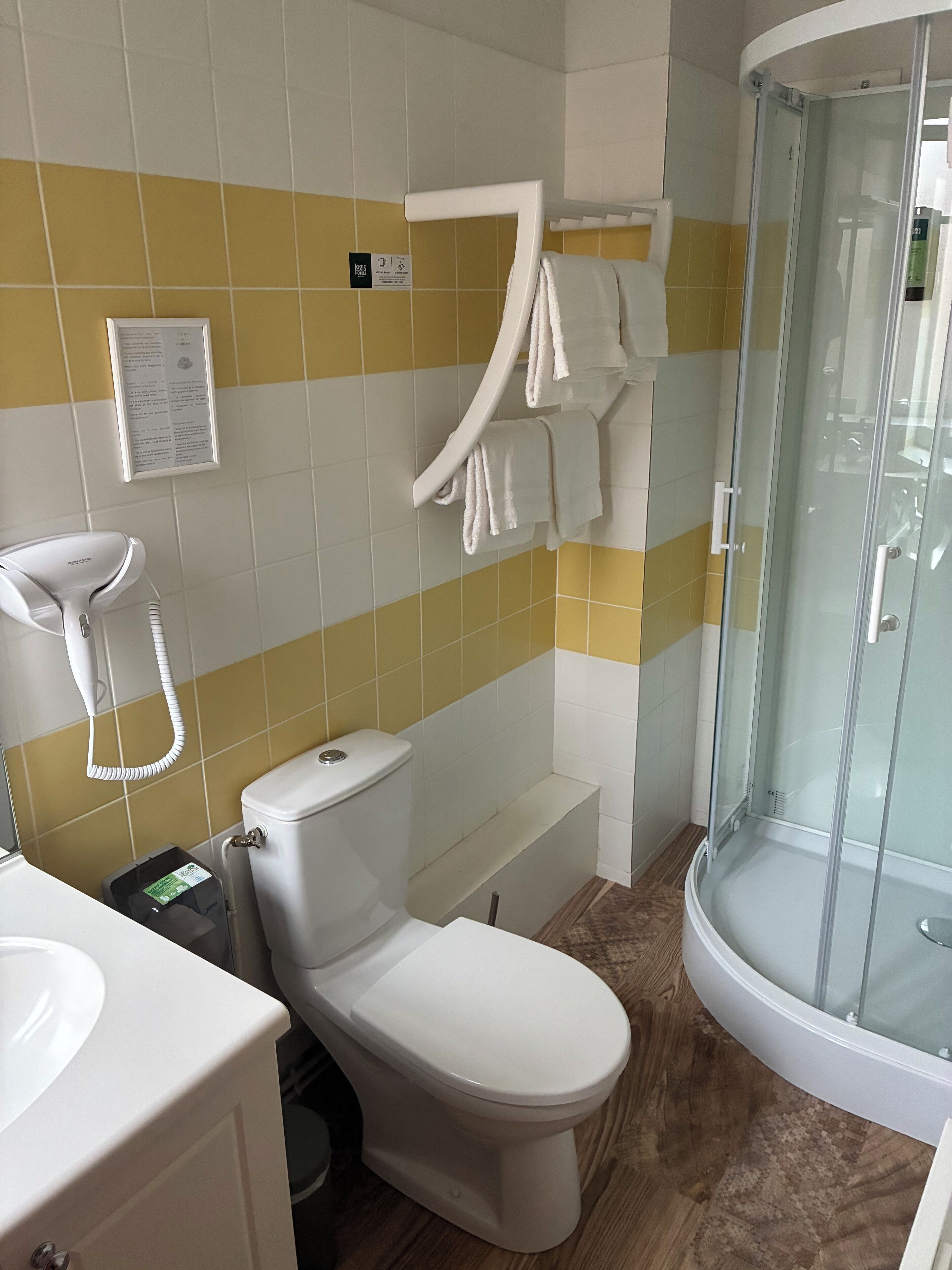 A bathroom with a toilet, sink, and shower. There is a towel rack with towels, a hand dryer, and a sign on the wall.