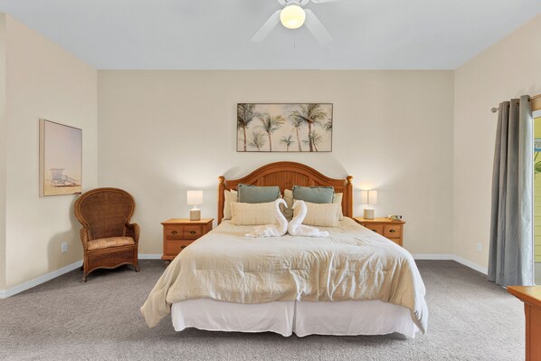 A neatly made bed with a wooden headboard, two bedside tables with lamps, a wicker chair, and a painting of palm trees on the wall.