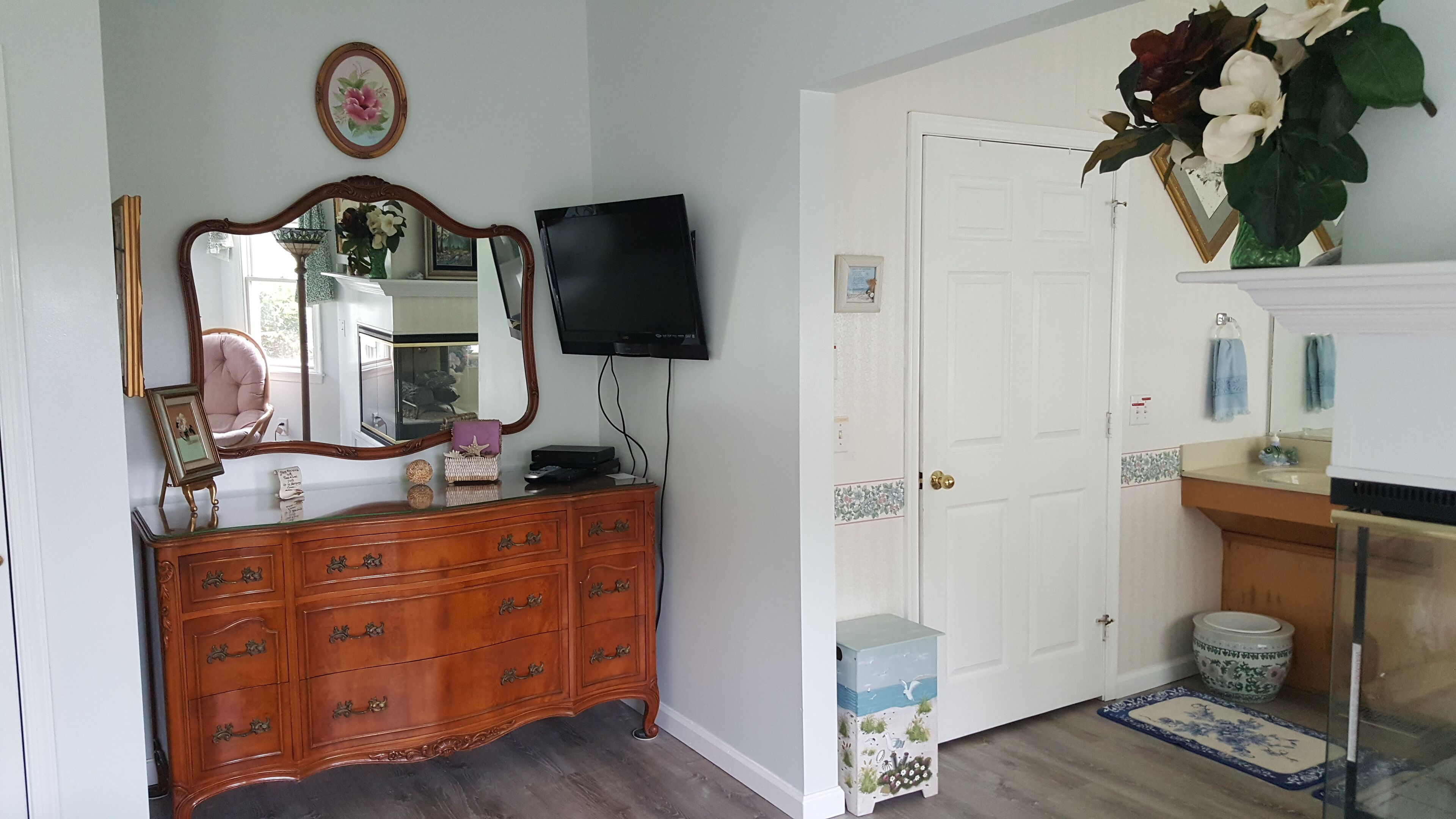 A room with a wooden dresser, a flat-screen TV, a mirror, a door, and a small trash bin.