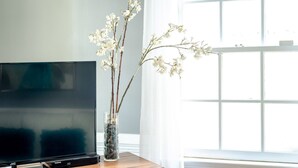 A flat-screen TV on a wooden table next to a window with white curtains.