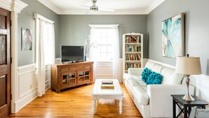 A living room with a white sofa, a coffee table, a TV on a wooden cabinet, a bookshelf, and a ceiling fan.
