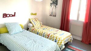 A bedroom with two beds, a wall with 'BOOM!' and 'Geral' decorations, a beach-themed picture, and a window with red curtains. - RENTAL HOUSE IN PEACE (Sibiril)