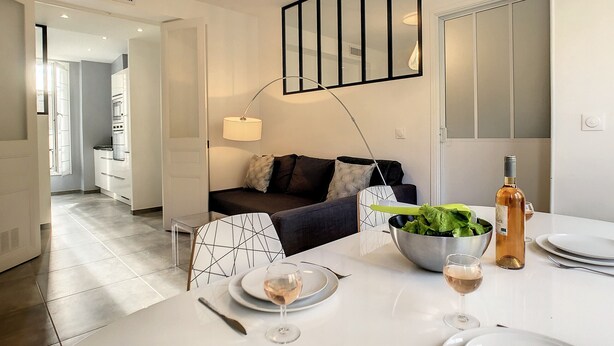 Une cuisine moderne avec des meubles blancs, un coin repas avec une table dressée pour deux personnes et un canapé agrémenté de coussins.