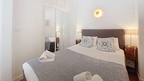 Una cama bien hecha con almohadones estampados, una lámpara de noche y una lámpara fijada a la pared.