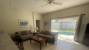 Un salón amplio con sofá, mesa de centro y televisor. Hay un ventanal corredero que da a una zona de piscina.
