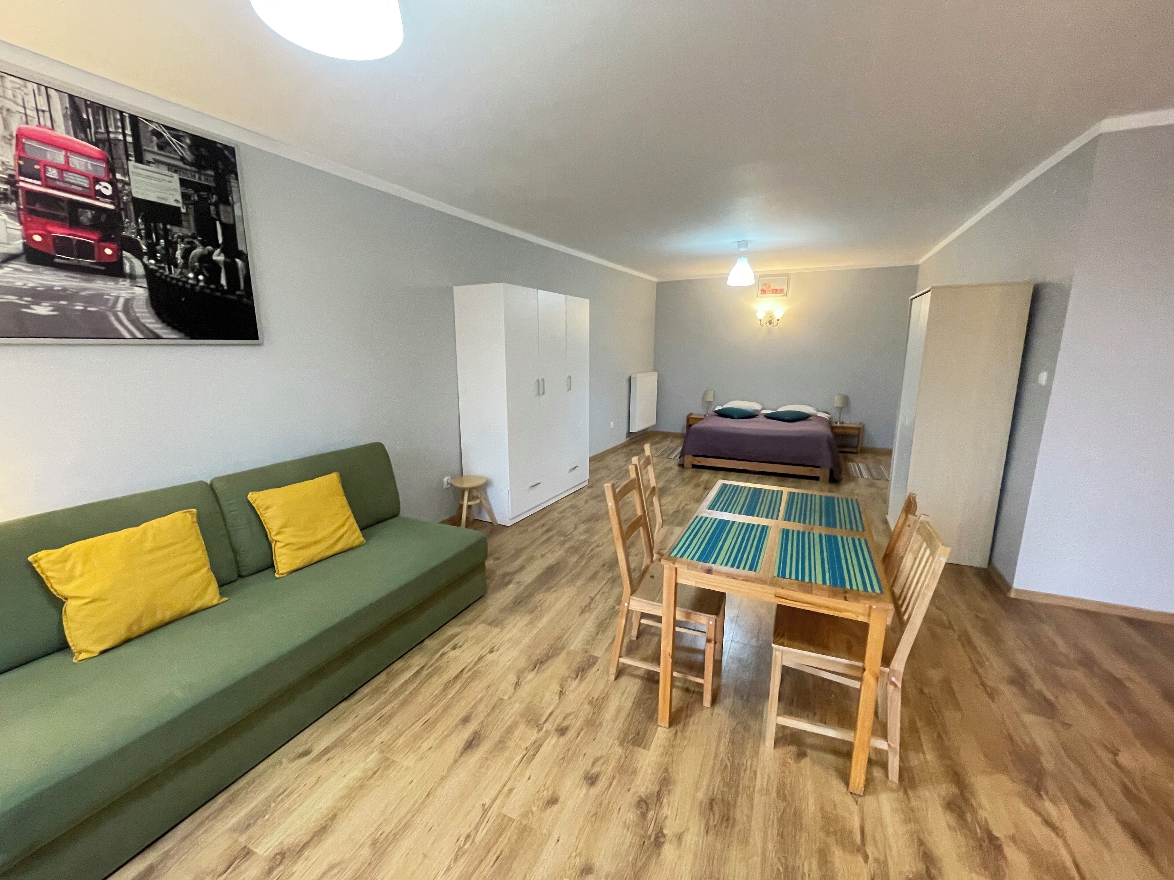 Una habitación con un sofá verde, una mesa de comedor de madera con sillas, una cama con colcha morada y un armario blanco.