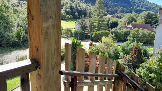 A wooden deck with a view of a rural landscape, including a house and a hill in the background. - Mountain view. Sleeps 4. Calm. Private parking. Shaded. Close to hiking trails. (La Bresse)