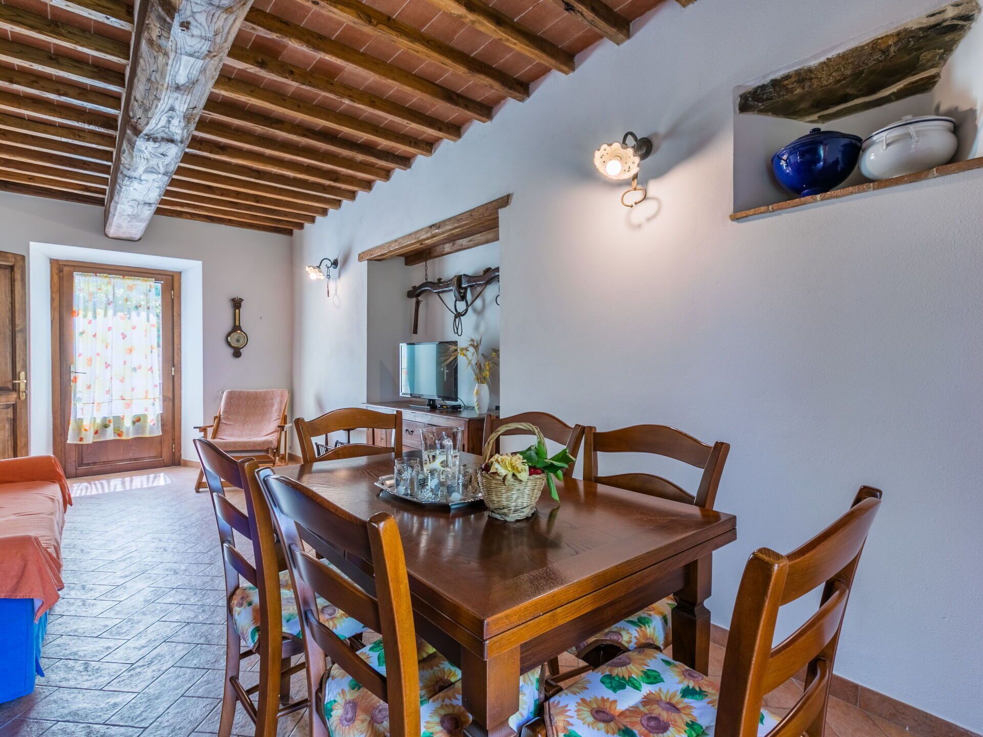 A dining area with a wooden table, chairs with floral upholstery, a television, and a shelf with decorative items.