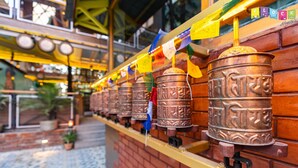 A row of prayer wheels mounted on a brick wall with a yellow railing. - Flock Kathmandu (Kathmandu)