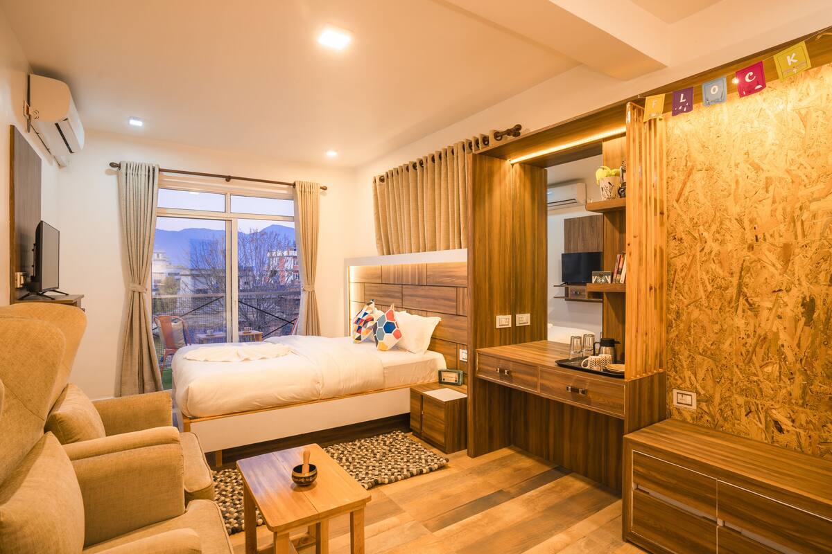 A modern hotel room with a bed, a sofa, a small table, a wooden cabinet, and a view of the outdoors.