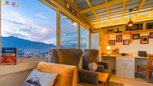 A cozy room with a view of a city and mountains, a comfortable chair, a desk with a laptop, and a shelf with various items. - Flock Kathmandu (Kathmandu)