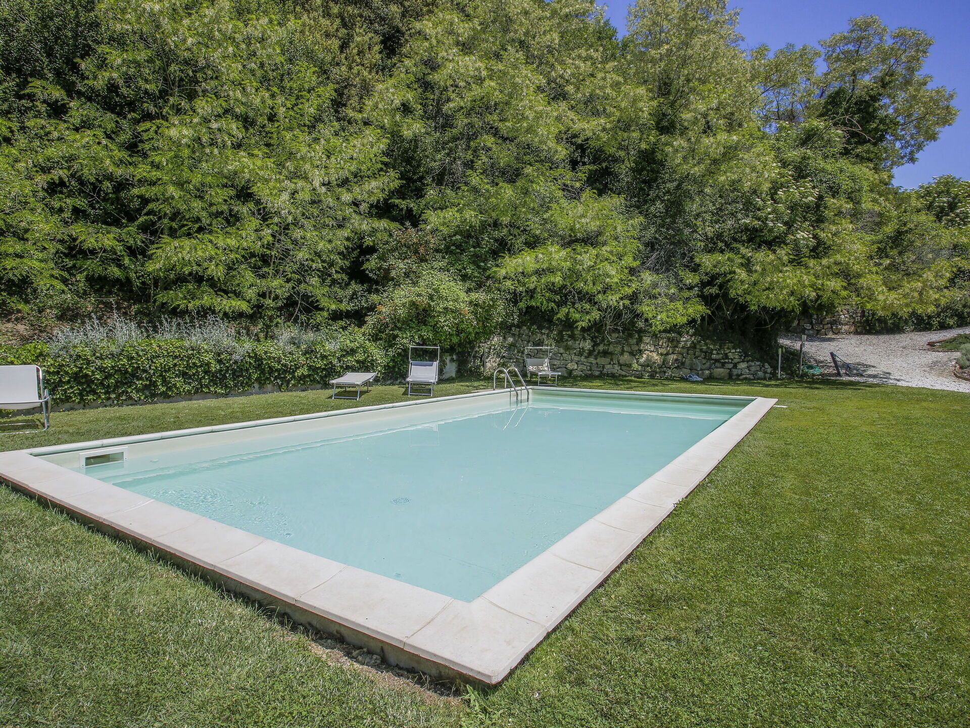 A rectangular swimming pool with a stone wall and a grassy lawn.