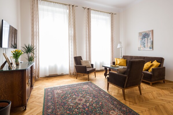Een woonkamer met houten vloer, een bank met gele kussens, een stoel, een salontafel, een televisie, een lamp en een schilderij aan de muur.