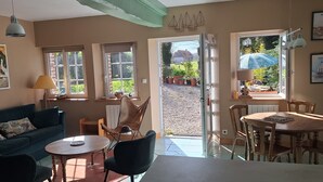 Ein gemütliches Wohnzimmer mit einer Couch, Sesseln, einem Tisch und einem Essbereich mit Stühlen und einem Tisch. Es gibt Fenster, eine Tür und einen Ausblick nach draußen.