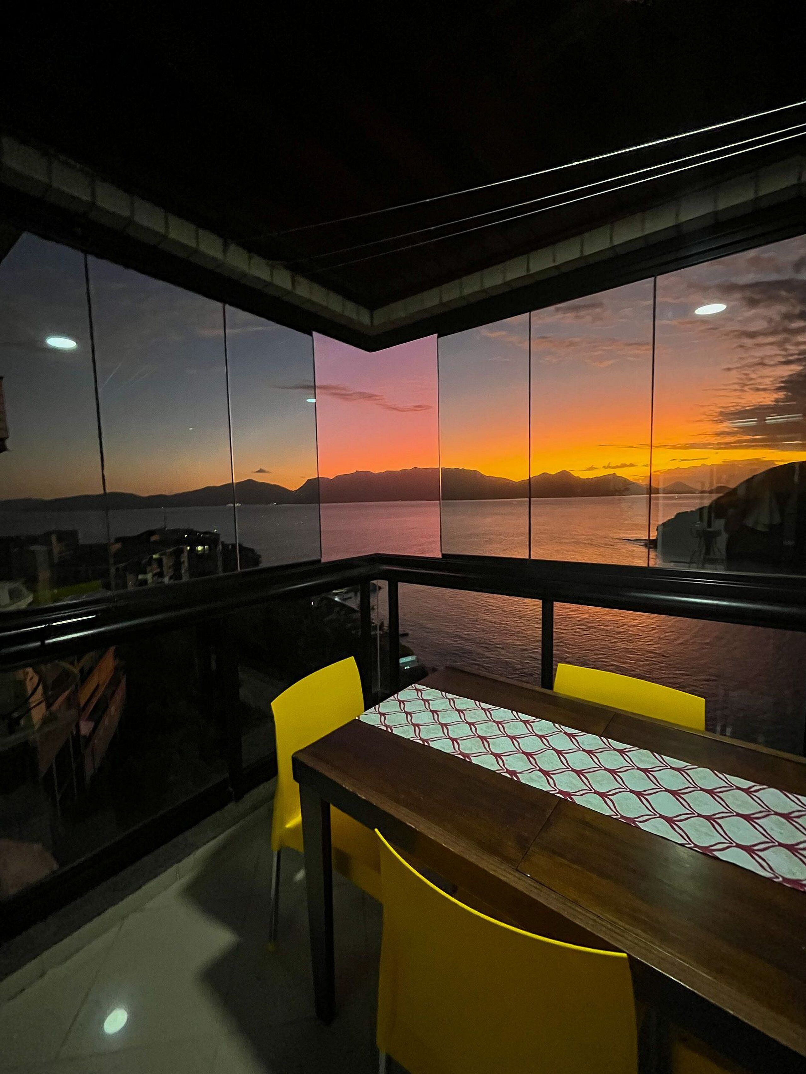 Un balcón con mesa y sillas, con vista a una puesta de sol sobre el agua y montañas.