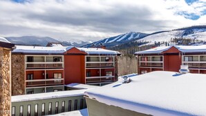 A row of red multi-story buildings with balconies, set against a snowy mountain backdrop. - Beautiful 2 bd/2 ba condo in the heart of town with all the amenities (Winter Park)