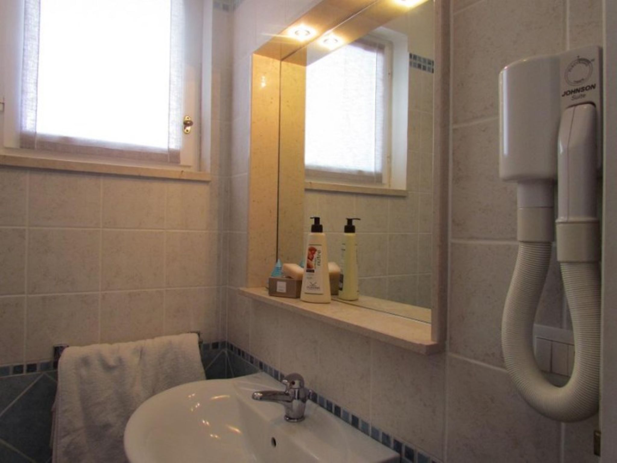 Un baño con lavamanos, espejo y perchero. Hay artículos de aseo en el lavamanos y un teléfono en la pared.