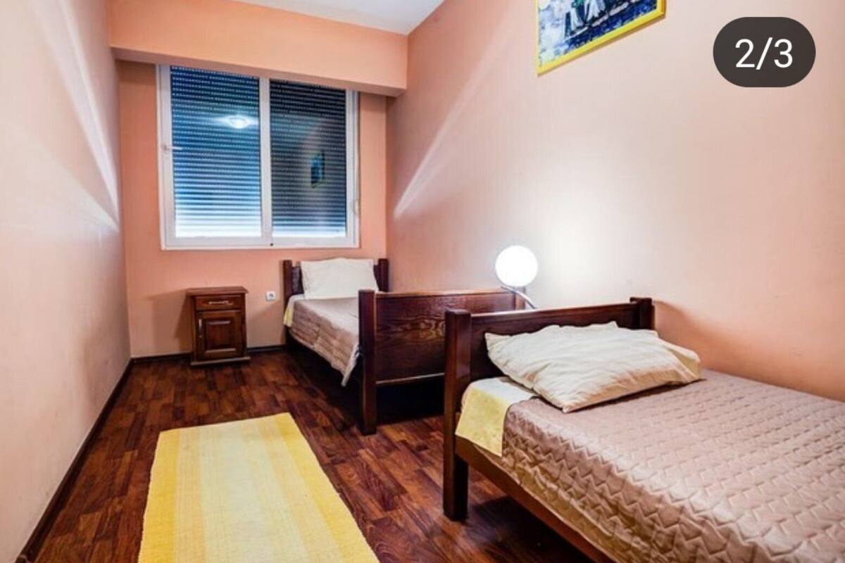 A small bedroom with two wooden beds, a bedside table, a lamp, and a window with blinds.