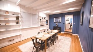 Uma sala de jantar com uma mesa de madeira posta para seis pessoas, rodeada por cadeiras. Há prateleiras brancas com diversos objetos à esquerda, e uma parede azul com um quadro emoldurado à direita.