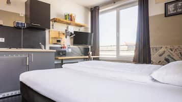 Ein modernes Hotelzimmer mit Bett, Schreibtisch, Flachbildfernseher, einer kleinen Küchenzeile und einem großen Fenster mit Vorhängen.