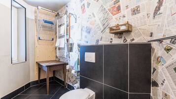 Een badkamer met een toilet, een houten bankje, een handdoekhouder en een met een krant beklede muur.