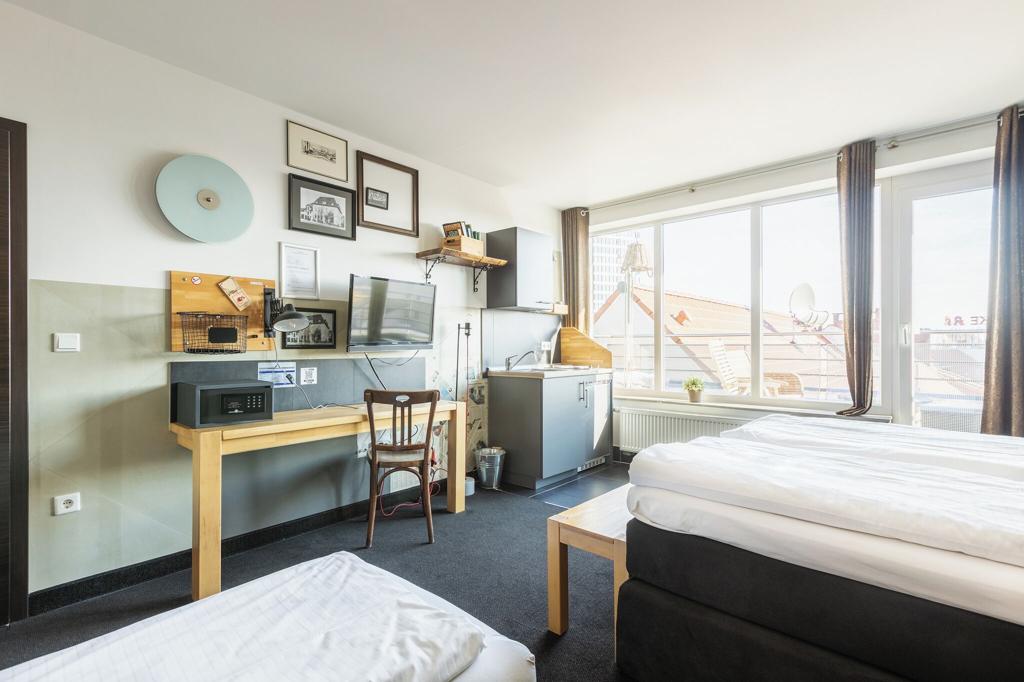A hotel room with a bed, desk, chair, kitchenette, and large windows.
