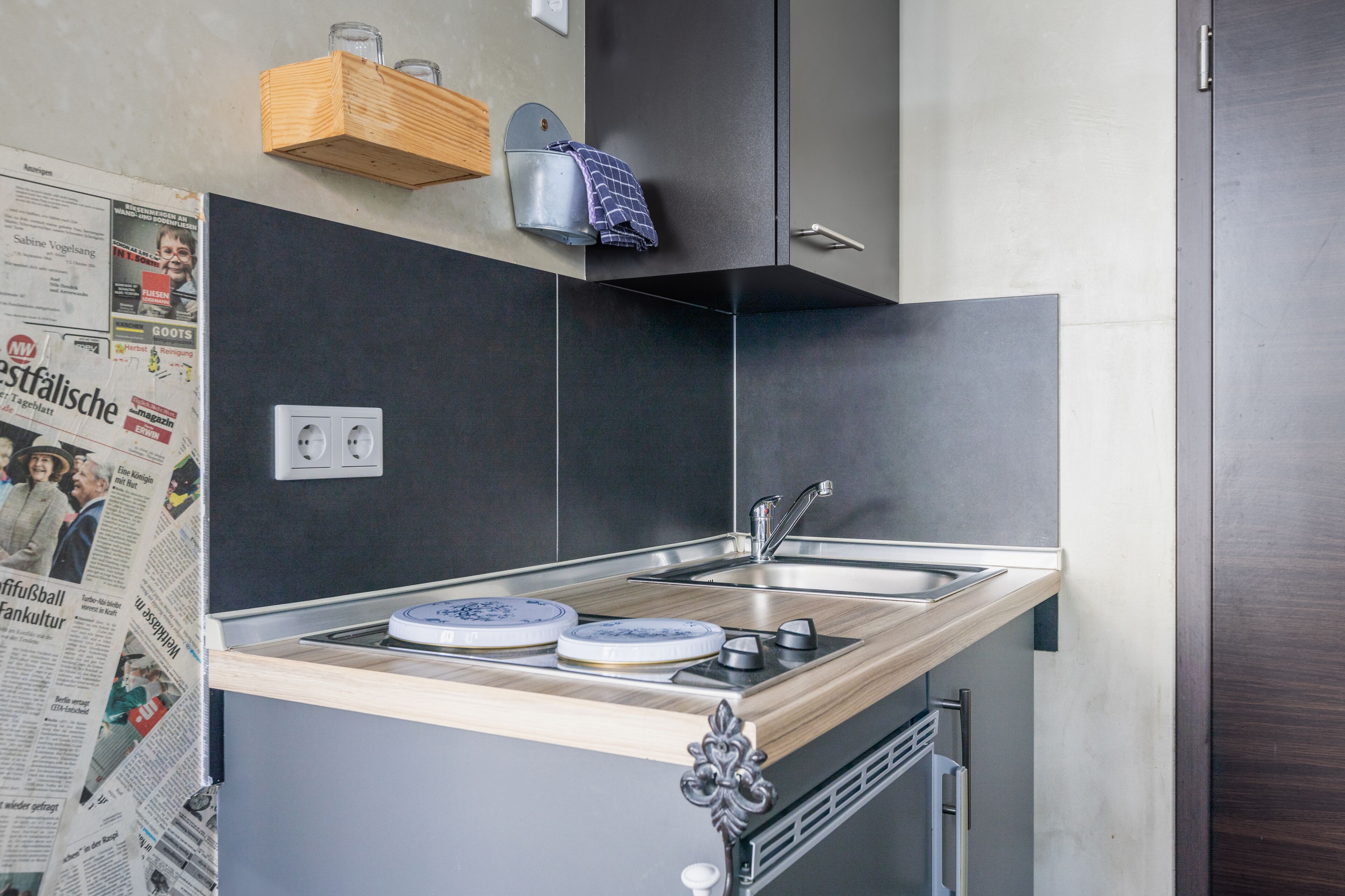 Cocina compacta con encimera de madera, un lavamanos y una estufa. Hay un periódico en la pared y una toalla colgada en un perchero.