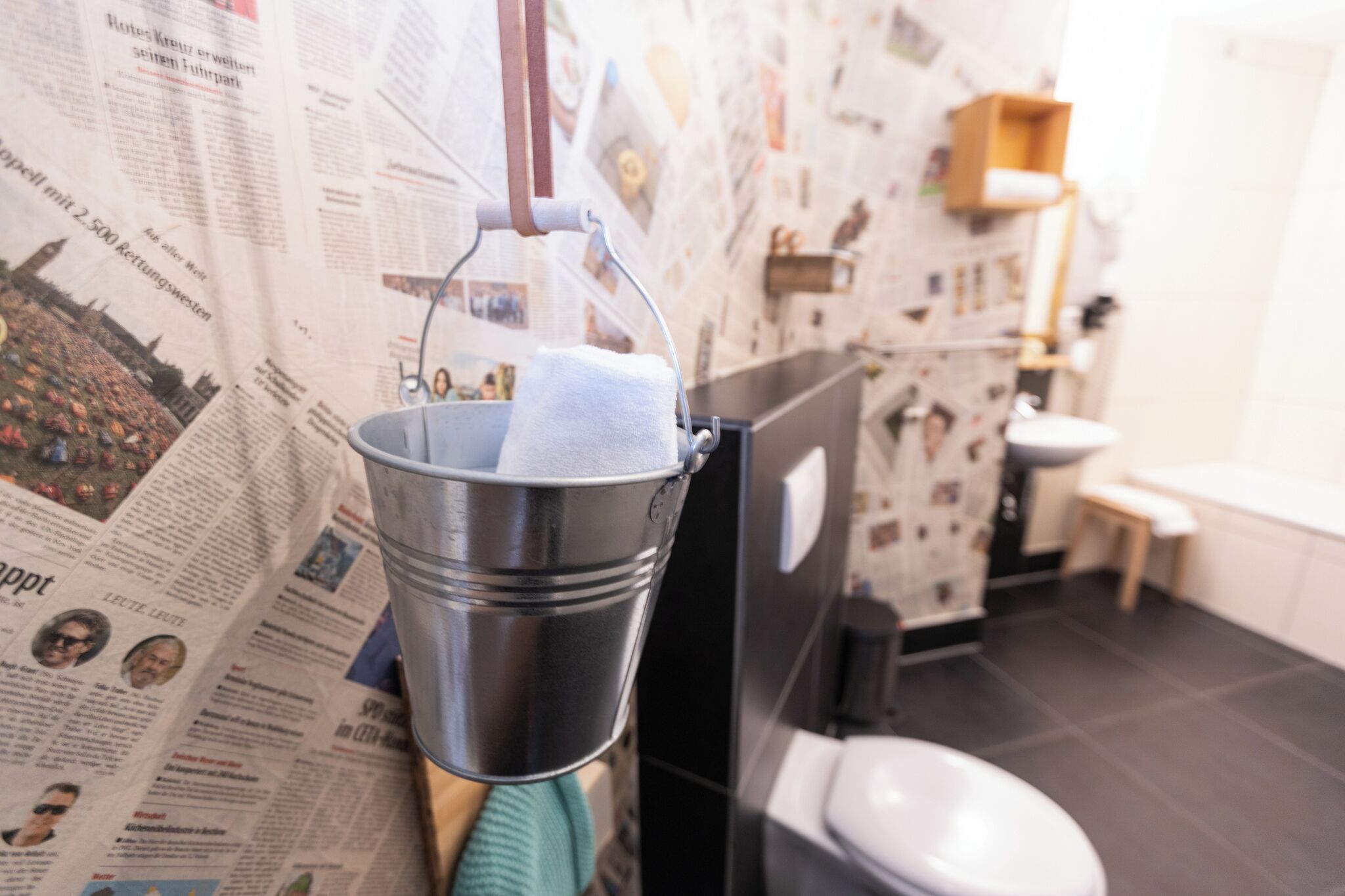 Ein Badezimmer mit einer an Zeitungspapier beklebten Wand, einem Metallkübel mit einem Handtuch, einer Toilette und einem Waschbecken.