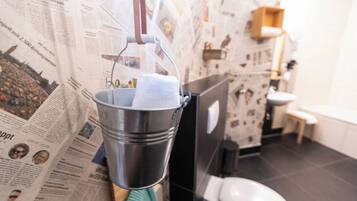 Ein Badezimmer mit einer an Zeitungspapier beklebten Wand, einem Metallkübel mit einem Handtuch, einer Toilette und einem Waschbecken.