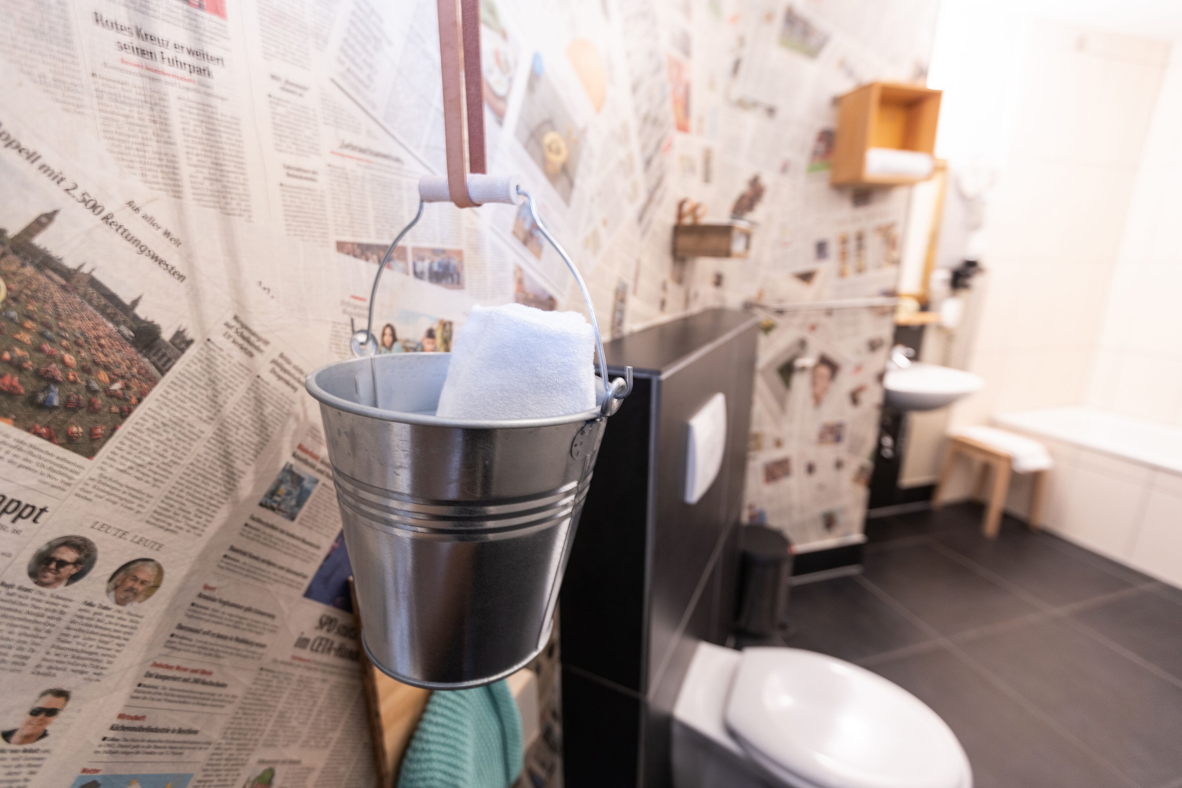A bathroom with a newspaper-covered wall, a metal bucket with a towel, a toilet, and a sink.