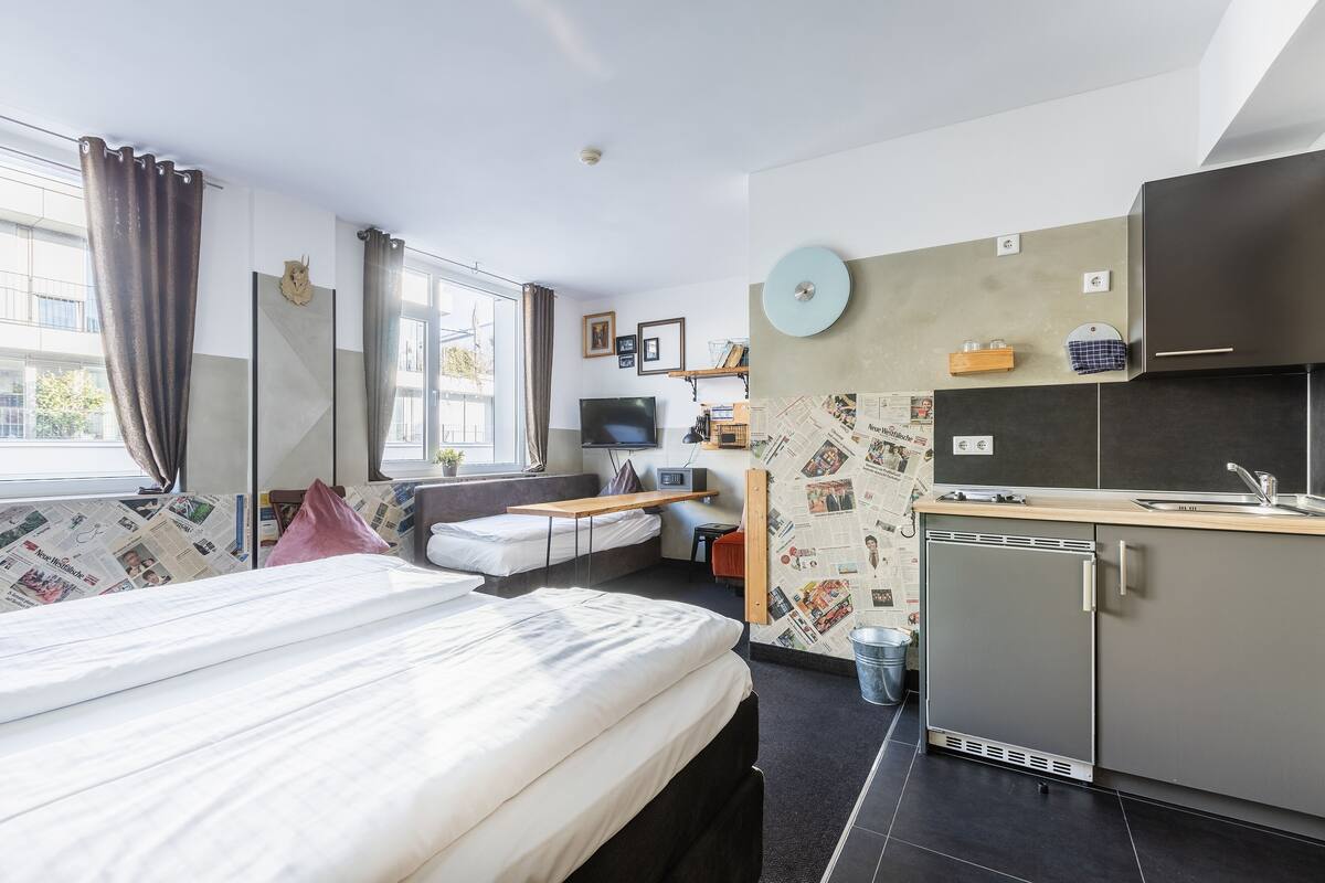 Een moderne hotelkamer met een bed, kitchenette, televisie en een raam met gordijnen.