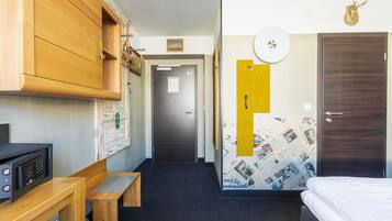 Une chambre d’hôtel comprenant un lit, un bureau en bois avec un micro-ondes, une armoire en bois, une porte avec un trou de serrure et un mur orné d’une carte tirée d’un journal.