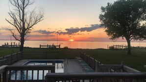 A deck with a view of the sunset over a body of water, surrounded by trees and a grassy area. - Tilghman | Pool | Private Dock | Pet Friendly (Tilghman)