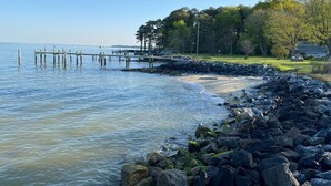 A coastal scene with a rocky shoreline, a wooden pier extending into calm waters, and a grassy area with trees and a house in the background. - Tilghman | Pool | Private Dock | Pet Friendly (Tilghman)